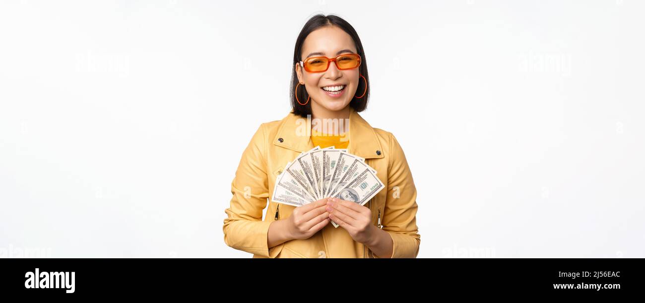 Ragazza asiatica sorridente di stile che tiene i soldi in contanti, mostrando i dollari e celebrando, in piedi su sfondo bianco Foto Stock