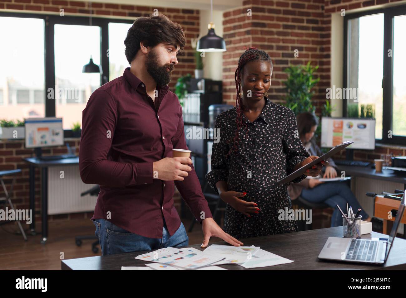 Donna d'affari afroamericana incinta che ha appunti che guardano i dati finanziari. Colleghi aziendali che rivedono le idee di marketing di progetto e la strategia aziendale mentre si trovano in ufficio di agenzia. Foto Stock