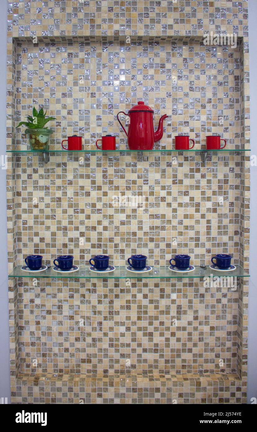 parete in piastrelle marrone bianco e giallo con decorazioni vintage, teiera e tazze, rosso e blu su una mensola di vetro e un vaso di vetro con una pianta Foto Stock