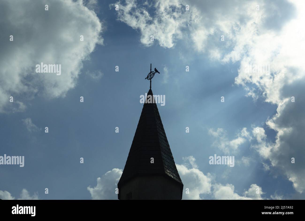 Un uccello perches sul weathervane su un campanile della chiesa. Le nuvole illuminate dal sole e i raggi solari si estendono verso l'esterno Foto Stock