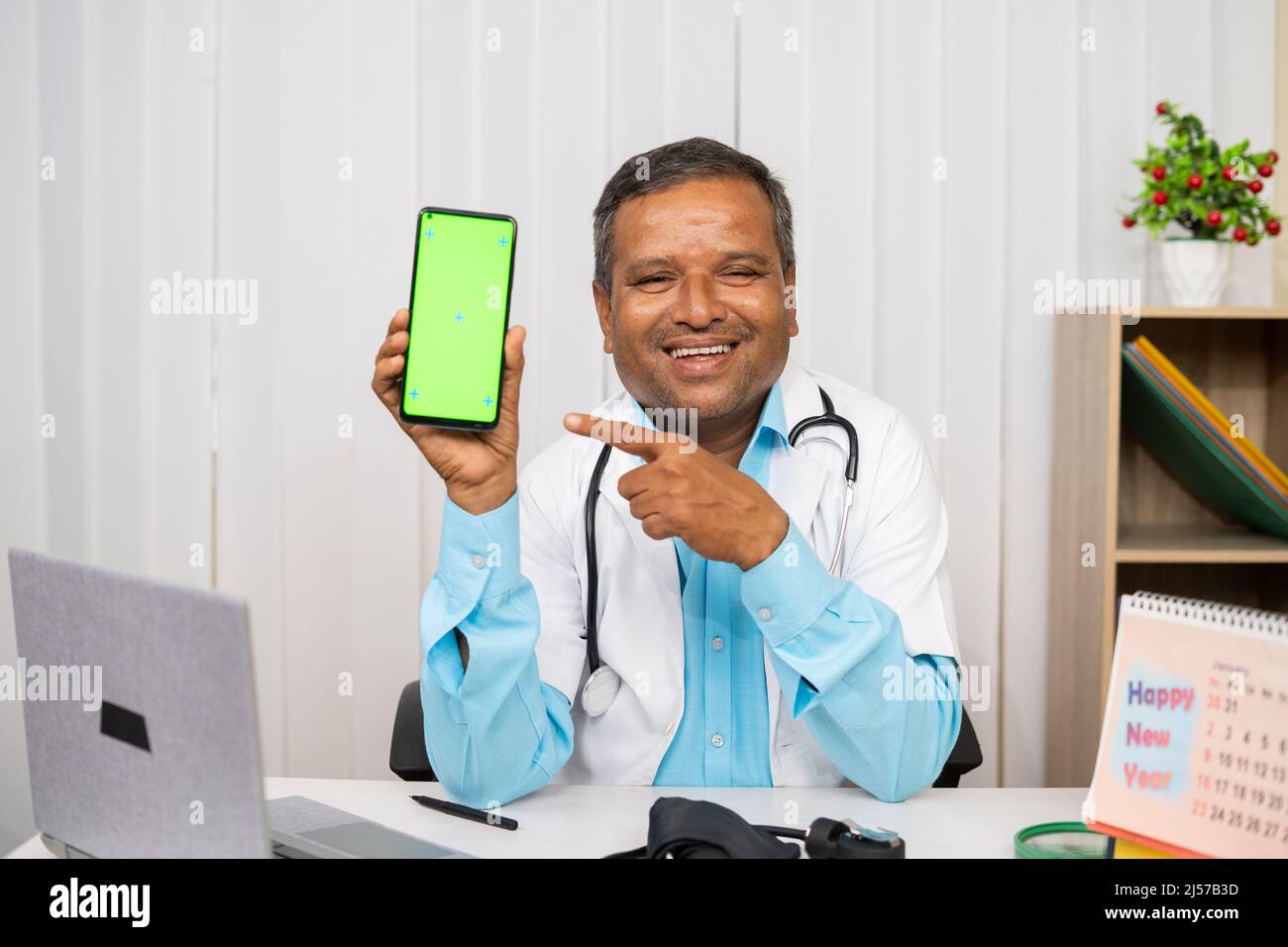 Sorridente medico che mostra il telefono cellulare con schermo verde puntando il dito mentre si guarda la macchina fotografica all'ospedale - concetto di salute, salute virtuale Foto Stock