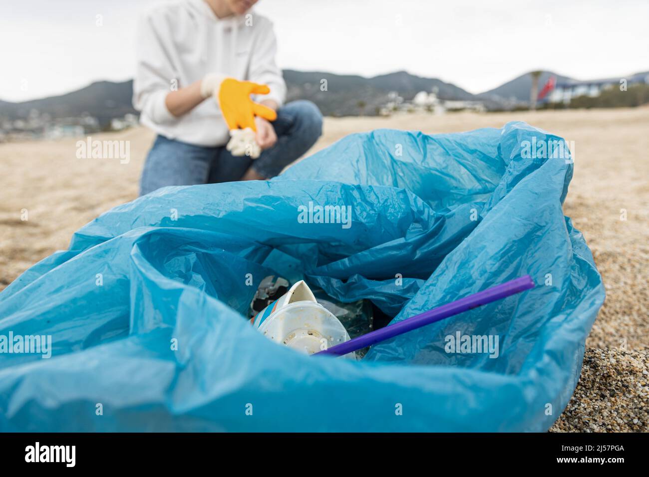 Una donna in guanti con una speciale borsa blu raccoglie rifiuti tra la sabbia lungo la costa. Il problema dell'inquinamento ambientale. Pulizia dei rifiuti Foto Stock