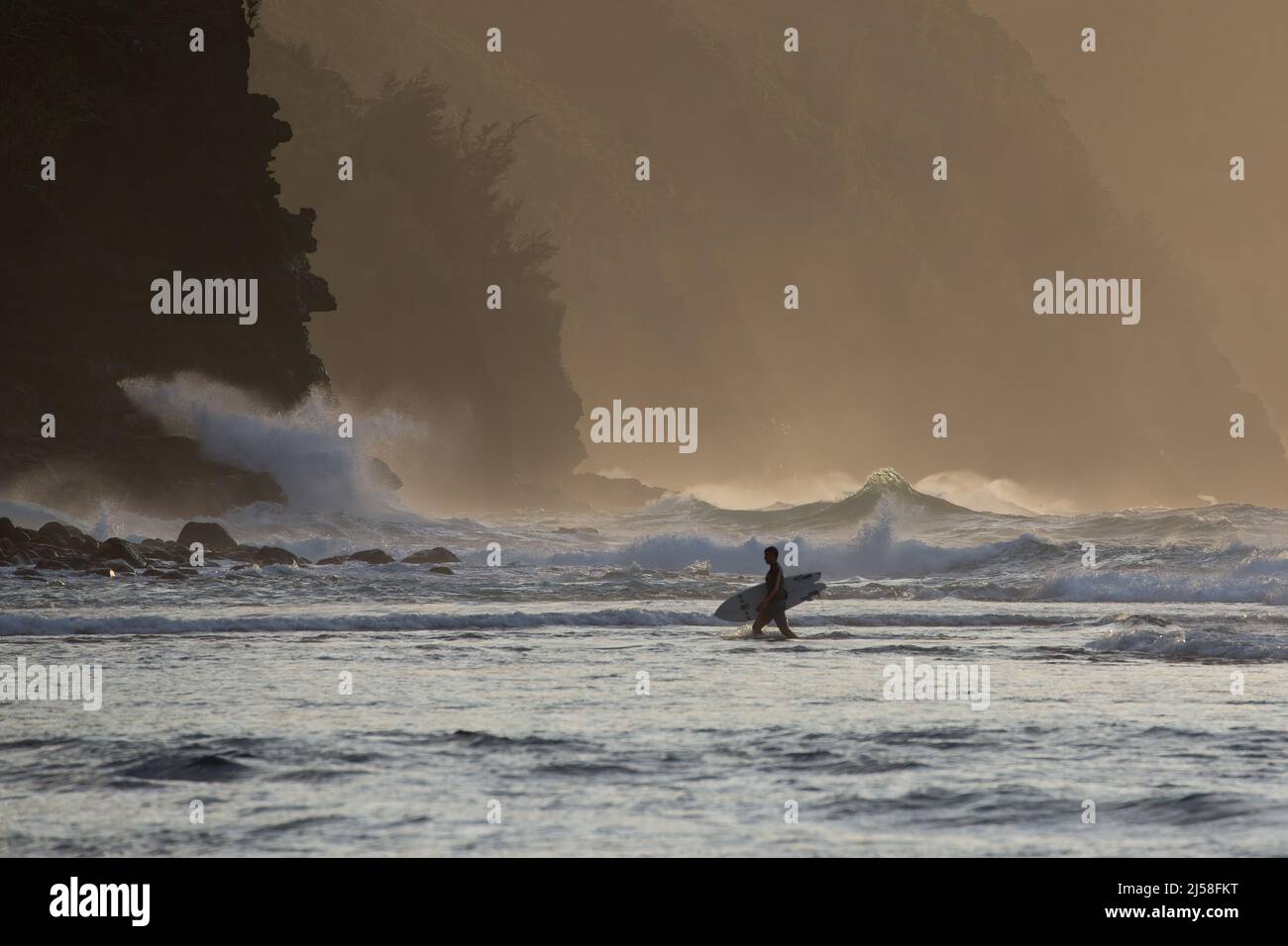 Un surfista entra a Ke'e Beach alla fine del giorno, mentre le onde si infrangono violentemente dietro le scogliere rocciose della costa di Na Pali a Kauai, Hawaii. Foto Stock