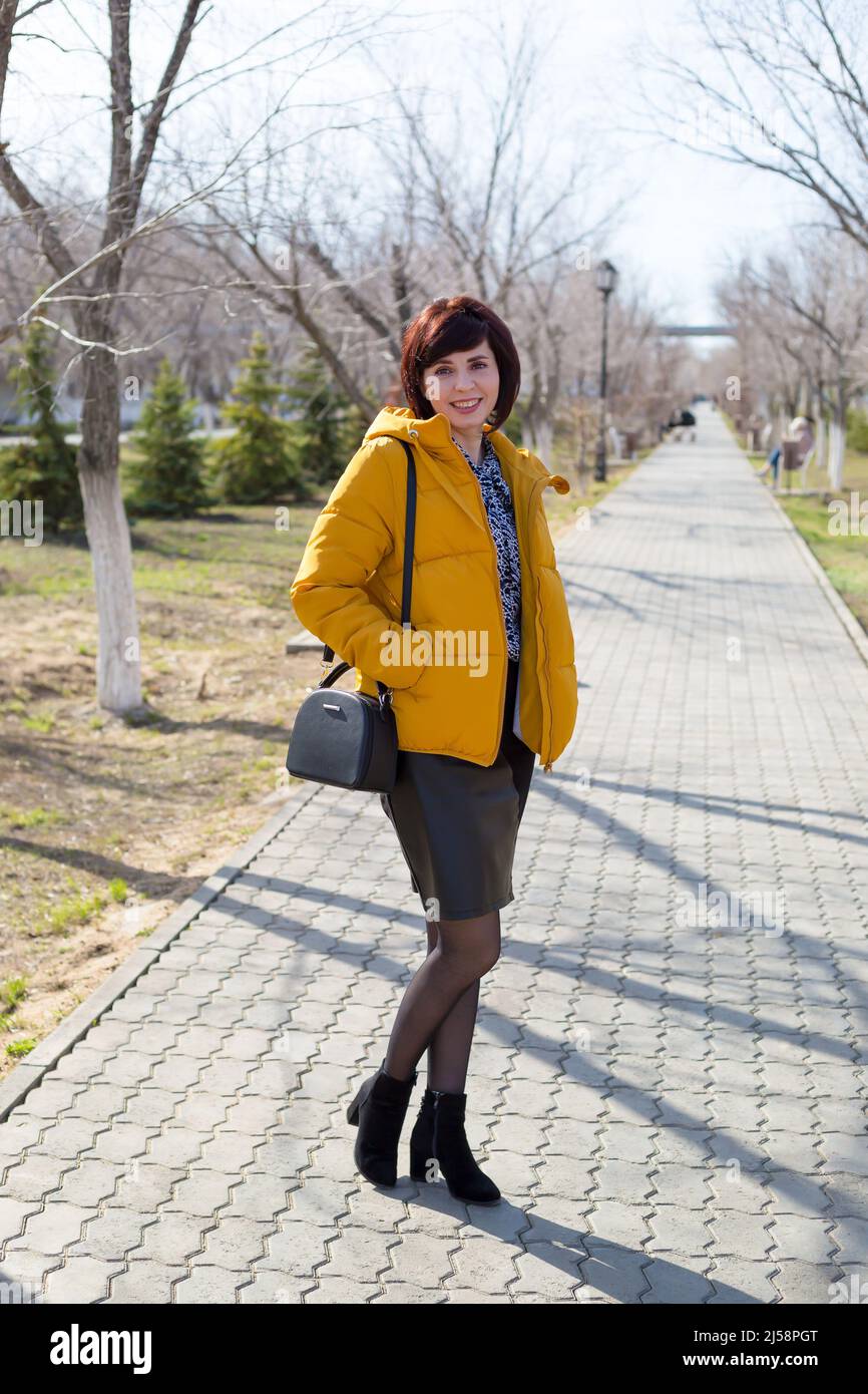 Una donna brunetta adulta di 40-44 anni in una giacca arancione brillante che cammina nel parco in una soleggiata giornata di primavera. Foto Stock