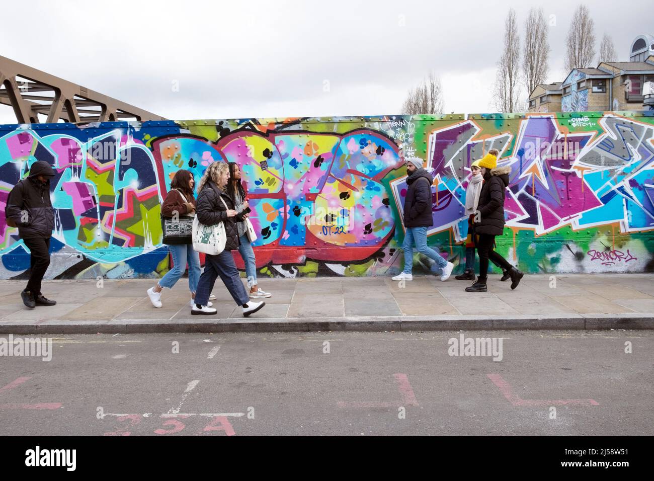 Persone pedoni a piedi da coloratissimi graffiti dipinti su hoardings in Brick Lane Shoreditch primavera Aprile 2022 Londra Inghilterra Regno Unito KATHY DEWITT Foto Stock