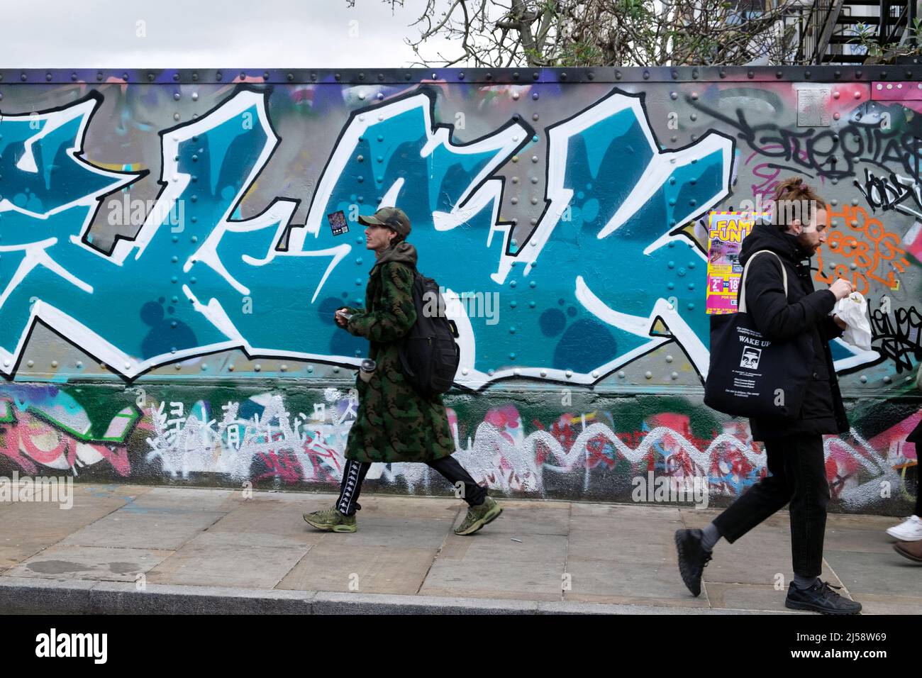Giovani uomini persone a piedi da coloratissimi graffiti pittura su hoardings in Brick Lane Shoreditch primavera Aprile 2022 Londra Inghilterra Regno Unito KATHY DEWITT Foto Stock