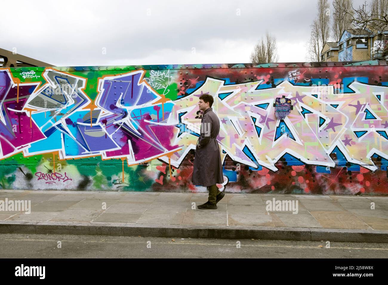 Uomo persona che indossa trench coat camminare con pittura colorata murale su Hoarding Brick Lane Shoreditch primavera aprile 2022 Londra Inghilterra Regno Unito KATHY DEWITT Foto Stock