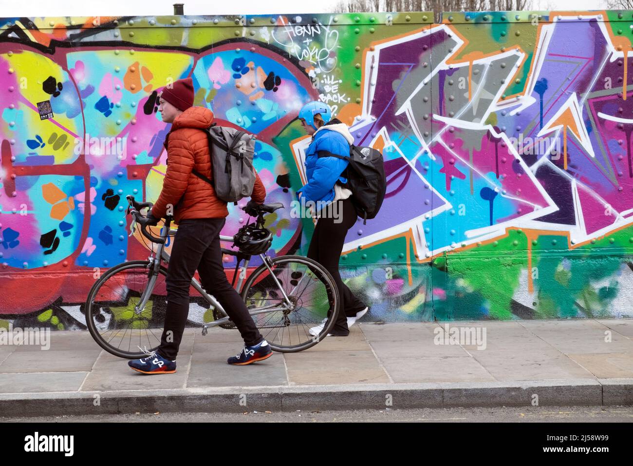 Cyclist coppia uomo bici a piedi da graffiti colorati pittura su hoardings in Brick Lane Shoreditch primavera Aprile 2022 Londra Inghilterra Regno Unito KATHY DEWITT Foto Stock
