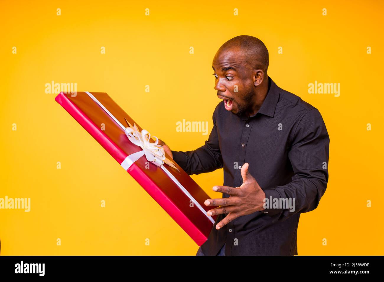 uomo ispanico americano in t-shirt nera ottenendo grande scatola regalo in studio sfondo giallo Foto Stock