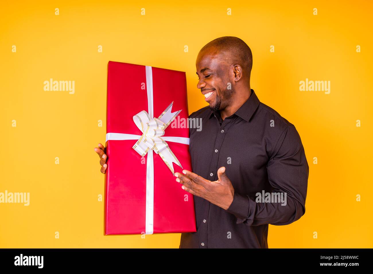 uomo ispanico americano in t-shirt nera ottenendo grande scatola regalo in studio sfondo giallo Foto Stock