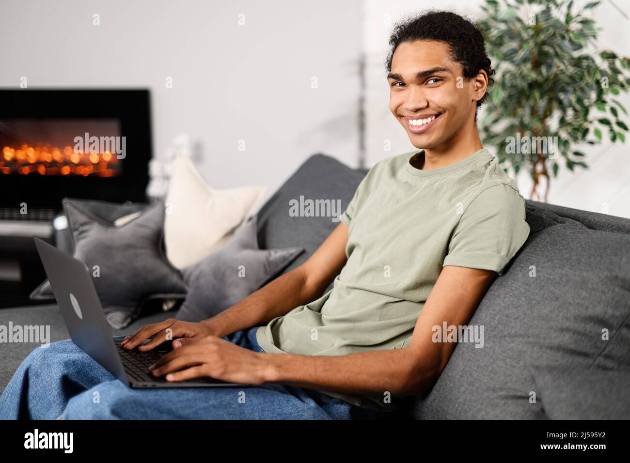Vista laterale dell'allegro uomo multirazziale indossando abbigliamento casual seduto sul divano e utilizzando un computer portatile in un moderno ufficio domestico. Felice imprenditore maschio digitando sulla tastiera e sorridendo alla fotocamera Foto Stock