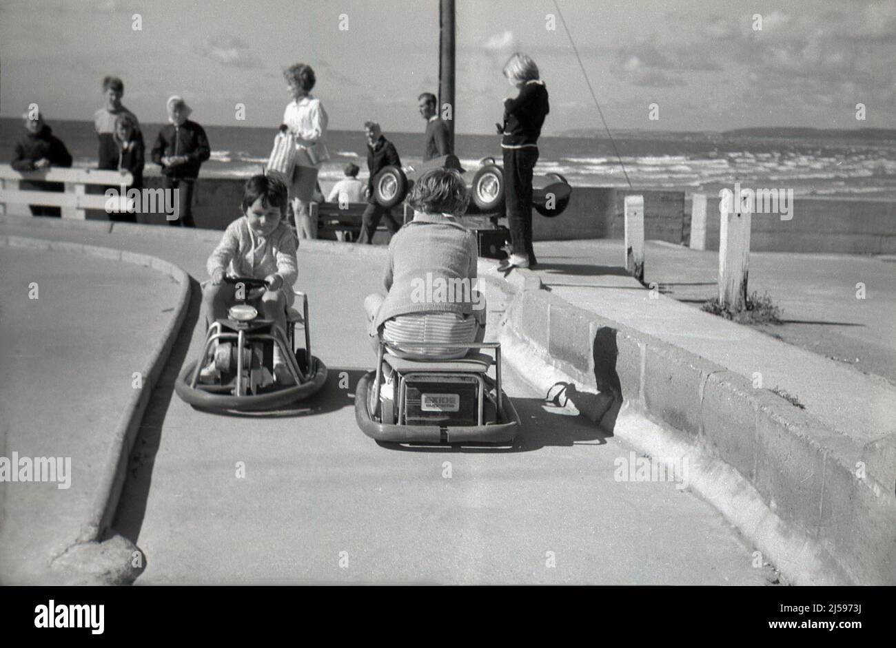 1960s, storico, al mare, due bambini piccoli che cavalcano su mini go-kart alimentati a batteria in una pista di cemento, Inghilterra, Regno Unito. Foto Stock