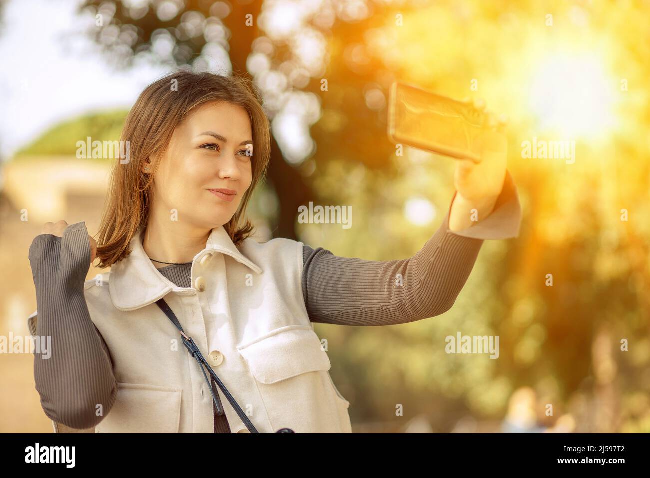 Allegra bella giovane donna prende un selfie su uno smartphone mentre si cammina all'aperto parco della città. Selfie da viaggio, blogger Foto Stock