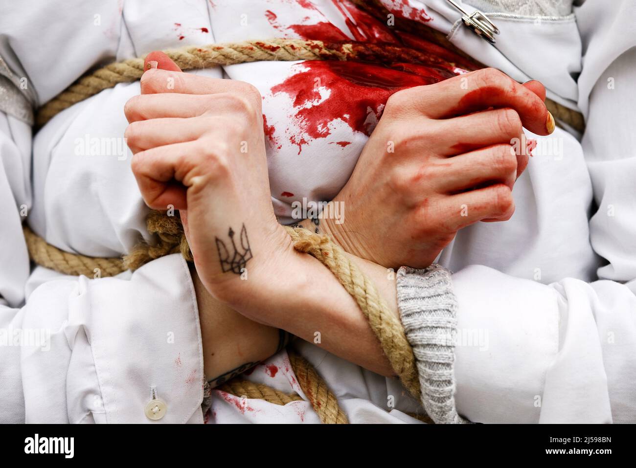 Berlino, Germania. 21st Apr 2022. Un partecipante ha un tatuaggio dello stemma ucraino in mano, come dimostra con le mani legate insieme ad altre donne in abiti bianchi con sangue falso su di loro e ha Unito le mani ad una dimostrazione contro la violenza contro le donne e le ragazze nella guerra russa Di aggressione all'Ucraina. Credit: Carsten Koall/dpa/Alamy Live News Foto Stock