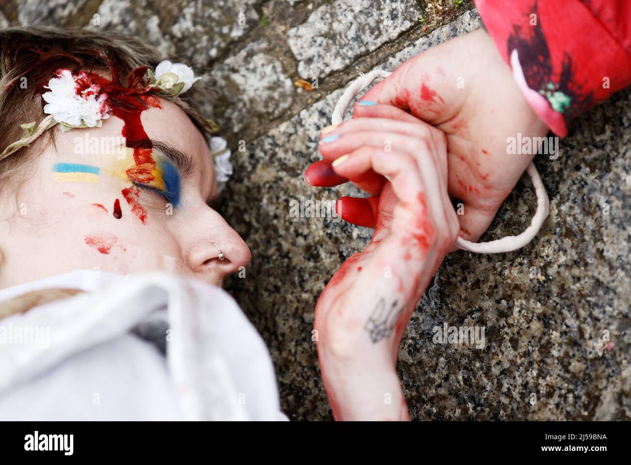 Berlino, Germania. 21st Apr 2022. Un partecipante con un tatuaggio dello stemma nazionale ucraino si trova a terra durante una manifestazione con altre donne in abiti bianchi con sangue artificiale su di loro e le mani legate insieme per protestare contro la violenza contro le donne e le ragazze in Guerra russa di aggressione all'Ucraina. Credit: Carsten Koall/dpa/Alamy Live News Foto Stock