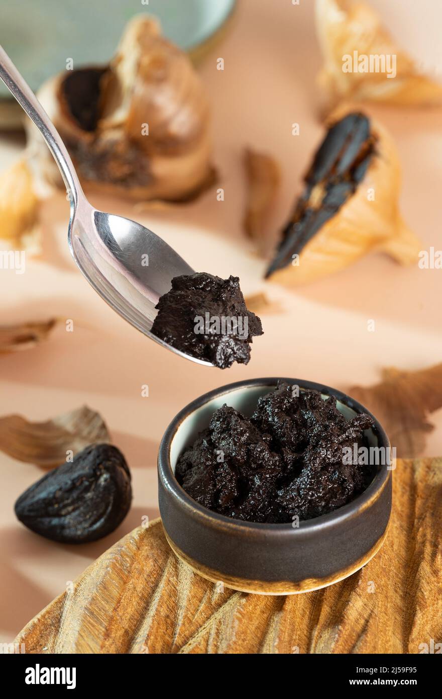 Cucchiaio con aioli e ciotola in ceramica argillosa con pasta d'aglio nera su un piatto di legno fatto a mano. Bulbo fermentato all'aglio sullo sfondo. Nutrizione sana Foto Stock
