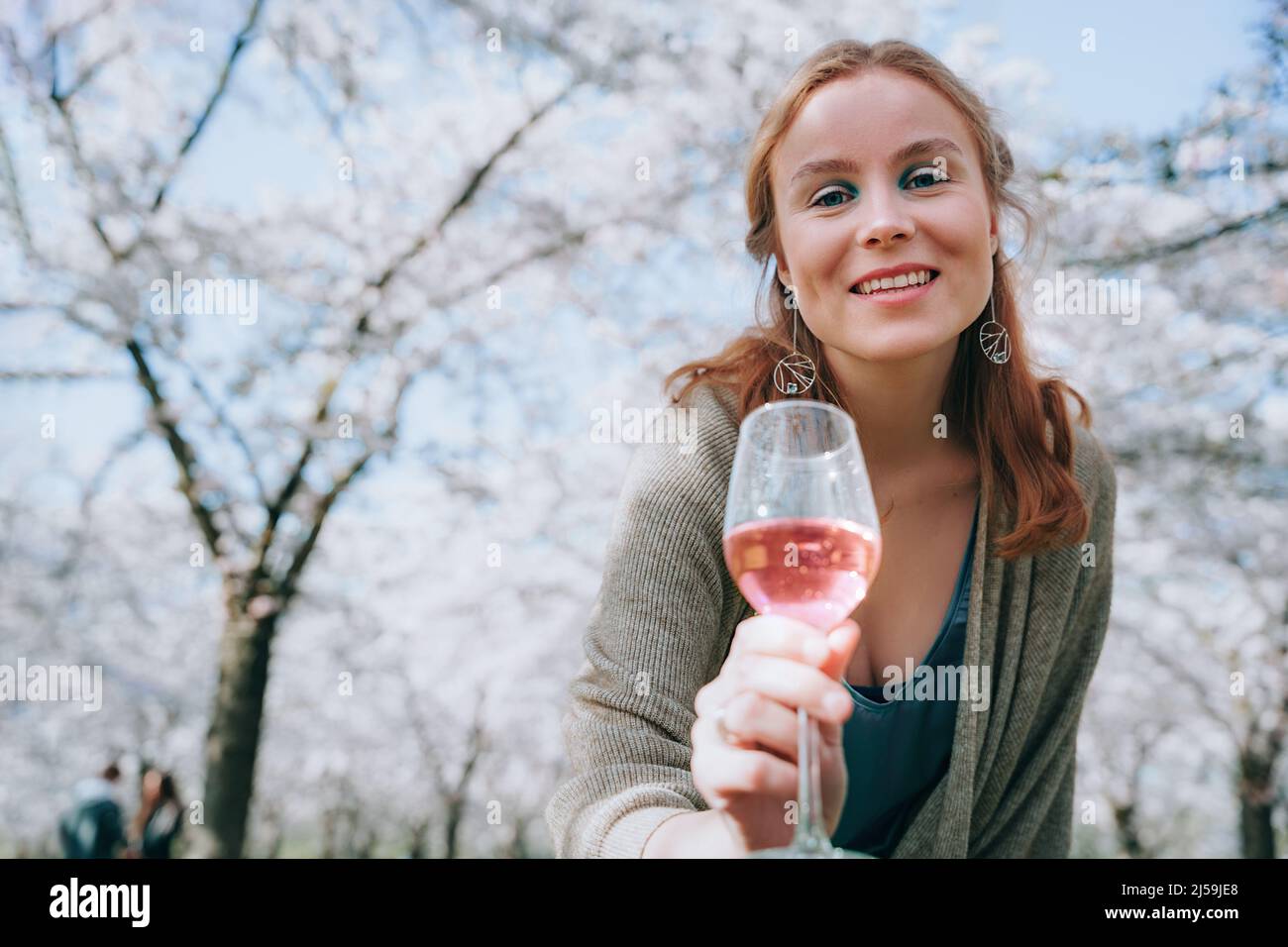 Ritratto di donna sorridente godendo la natura in primavera. Picnic sotto alberi di ciliegio fiorito. Donna che ama la vita, bere vino, offrire un bicchiere di vino a. Foto Stock