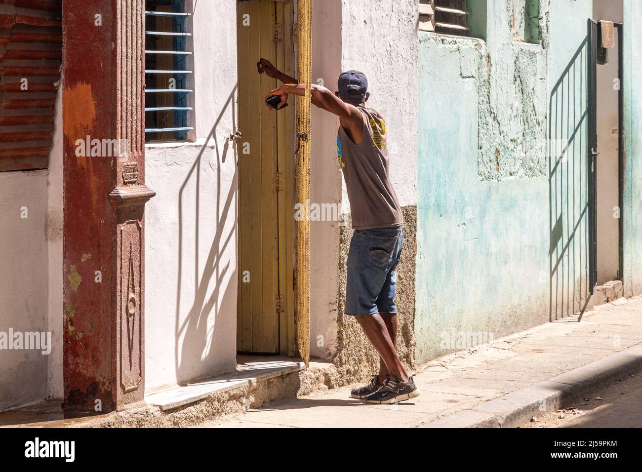 Un sottile uomo cubano afro-caraibico sta aspettando alla porta di una casa. È appoggiato su una griglia metallica protettiva che è a metà aperta. Stile di vita di persone reali io Foto Stock