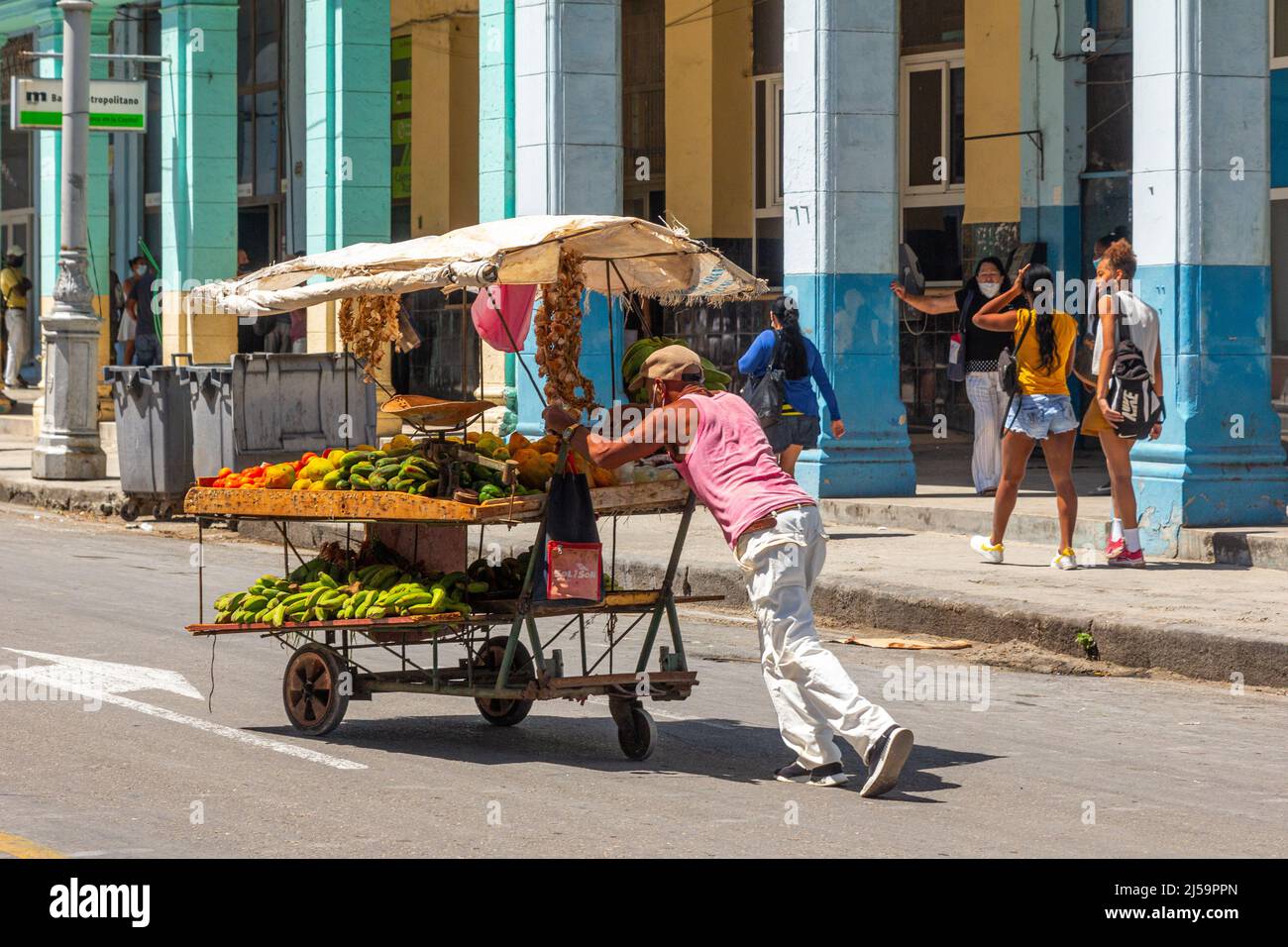 Un uomo cubano sottile spinge un carrello di frutta e verdura in una strada della capitale. Altre persone incidentali sono viste in background dal tempo Foto Stock