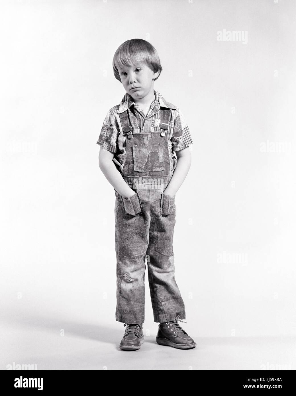 1970S RAGAZZO IN PIEDI INDOSSANDO IL DENIM PATCHED STRESSATO BIB TUTE LE MANI NELLE SUE TASCHE SAD VISO ESPRESSIONE GUARDANDO LA FOTOCAMERA - J13782 HAR001 HARS HOME VITA TASCHE COPIA SPAZIO TUTE A TUTTA LUNGHEZZA MASCHI DENIM ESPRESSIONI DISTURBATE B&W INTERESSATO TRISTEZZA OCCHIO CONTATTO ANGOSCIATO IL SUO UMORE POOUTING CONCETTUALE GUM POUT BIB GIOVANI MISERABILE PATCHED NERO E BIANCO ETNIA CAUCASICA HAR001 VECCHIO STILE Foto Stock