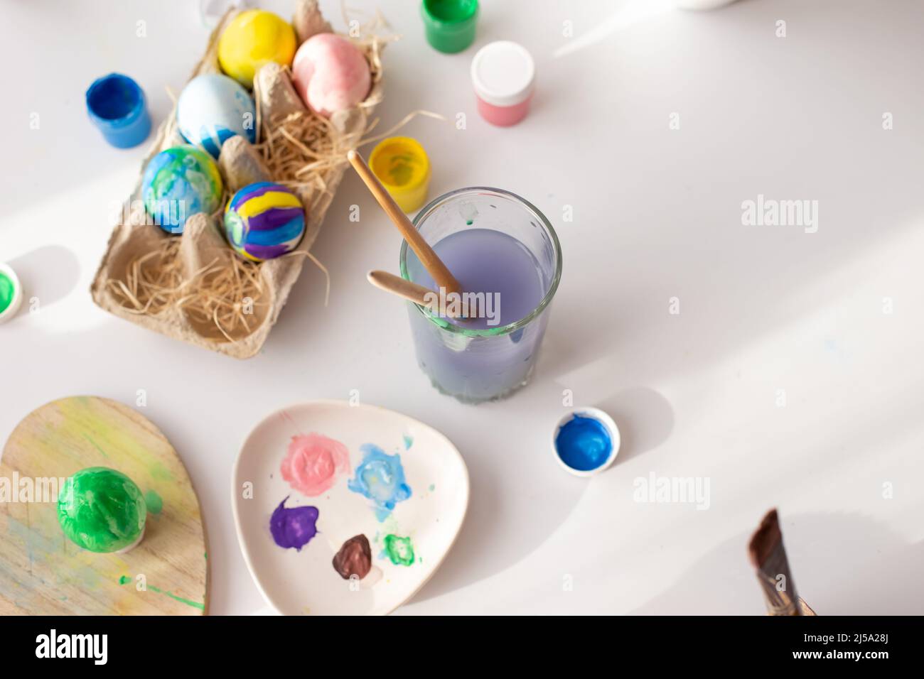 Su un tavolo bianco, alla luce del giorno, ci sono pennelli in un bicchiere d'acqua, uova multicolore in un vassoio, vasetti di vernice Foto Stock