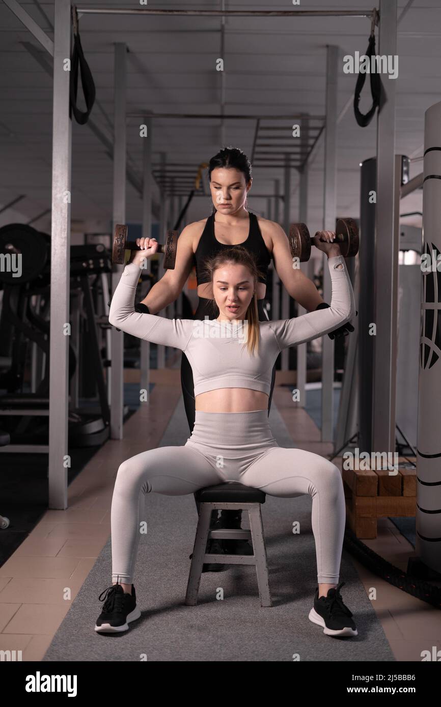 Shot di una giovane donna muscolare sollevamento manubri in palestra, allenamento con un personal trainer. La signora sta pompando i suoi muscoli della spalla con un enigma Foto Stock Shot di una giovane donna muscolare sollevamento manubri in palestra, allenamento con un personal trainer. La signora sta pompando i suoi muscoli della spalla con un enigma Foto Stock