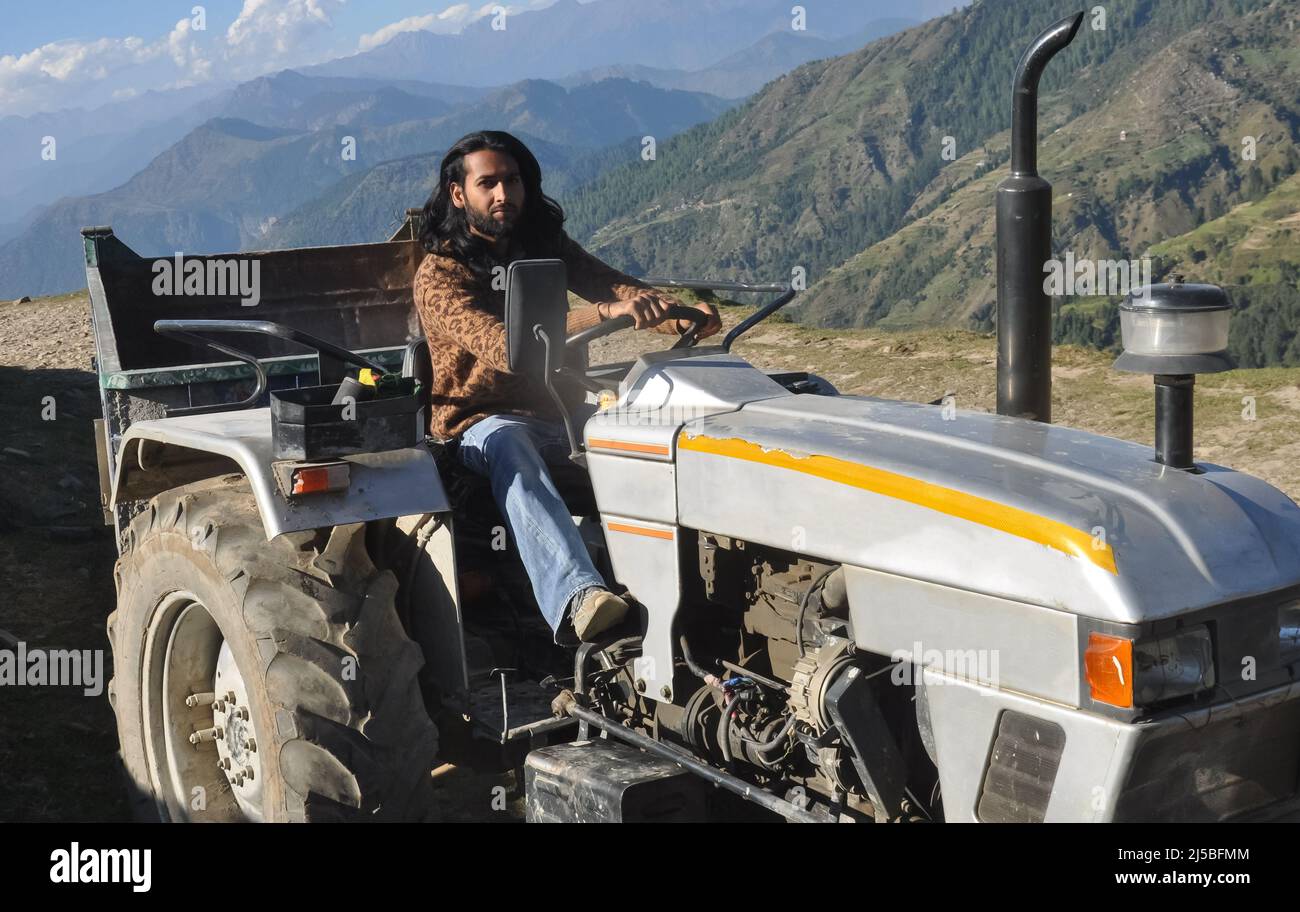 Un uomo dal bearded e dai capelli lunghi dell'asia del sud che guarda la macchina fotografica mentre guida un trattore vecchio in zona collinare Foto Stock