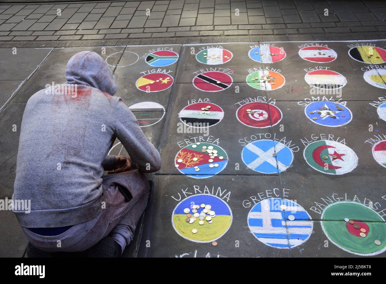 Un artista di marciapiede, che indossa una tuta grigia, disegna immagini di bandiere o bandiere di molti paesi del mondo su un marciapiede pubblico nel centro di Manchester, Inghilterra, Regno Unito, Isole britanniche. I passanti lasciano le monete sulle immagini della bandiera che amano. Foto Stock