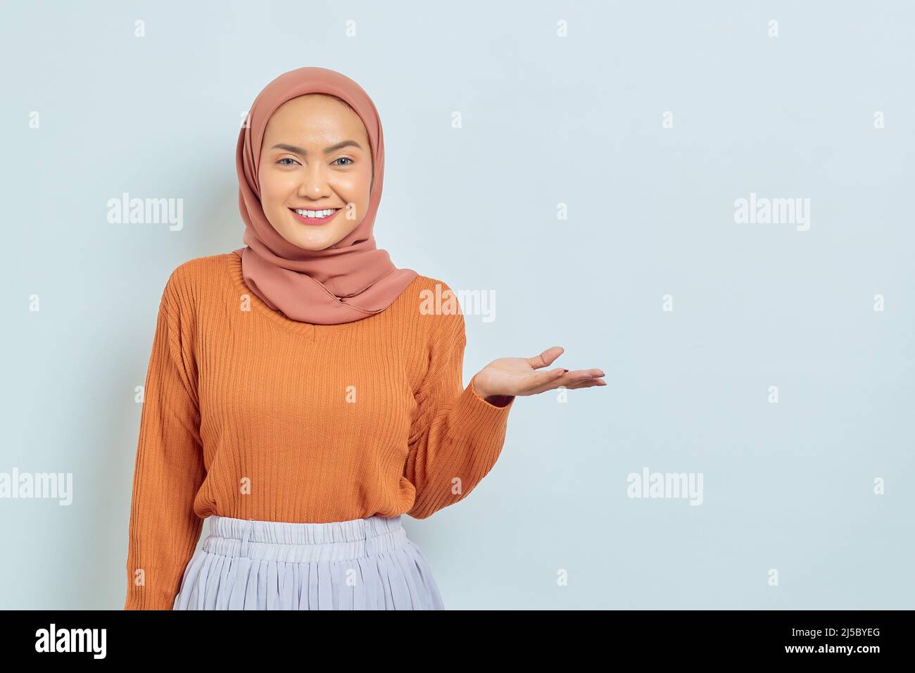 Sorridendo bella donna asiatica in maglione marrone e hijab puntando a copiare spazio con palme, mostrando prodotti pubblicitari e guardando con fiducia ca Foto Stock