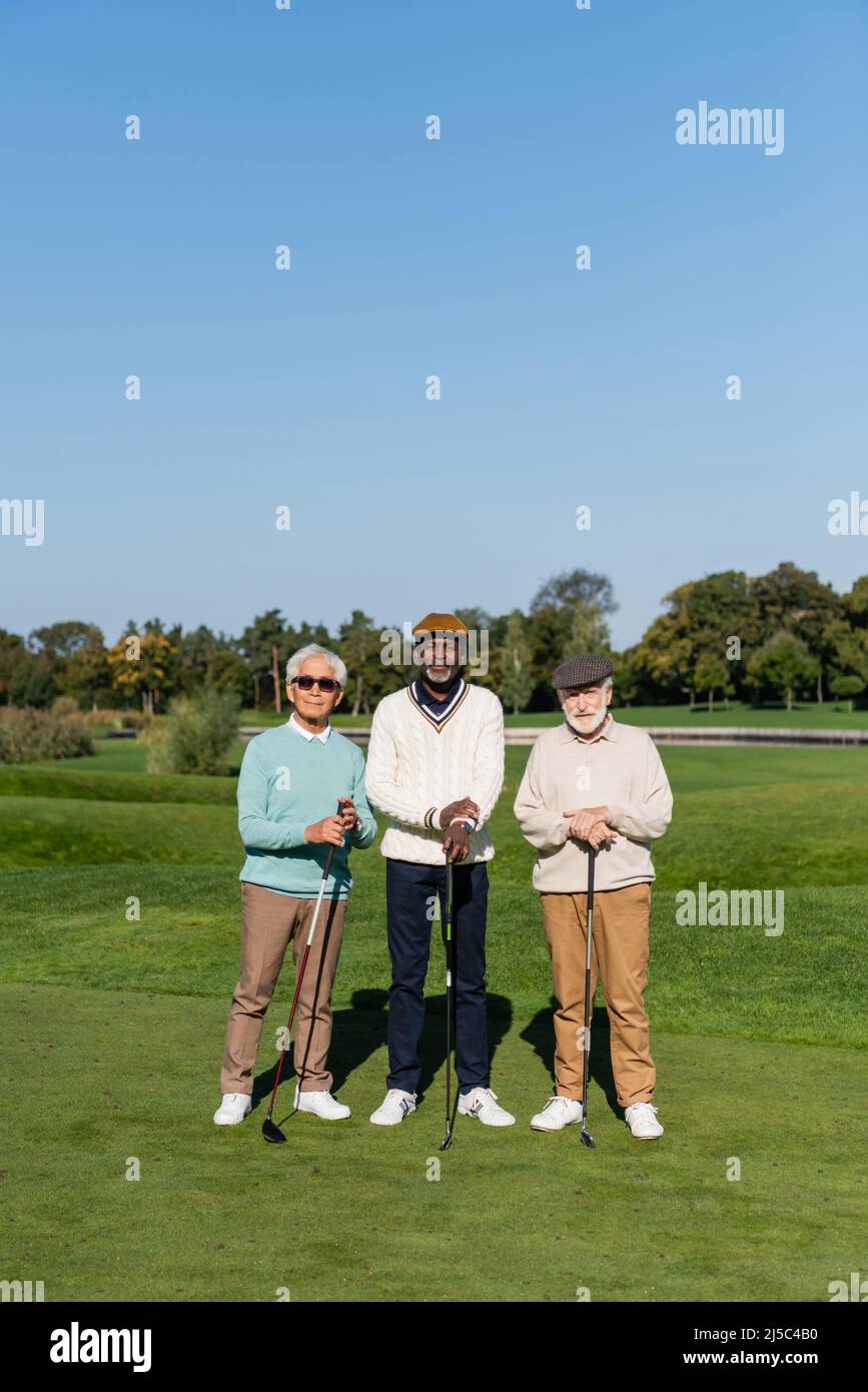uomo asiatico in occhiali da sole in piedi vicino ad amici multietnici anziani in calotte piatte con club di golf Foto Stock