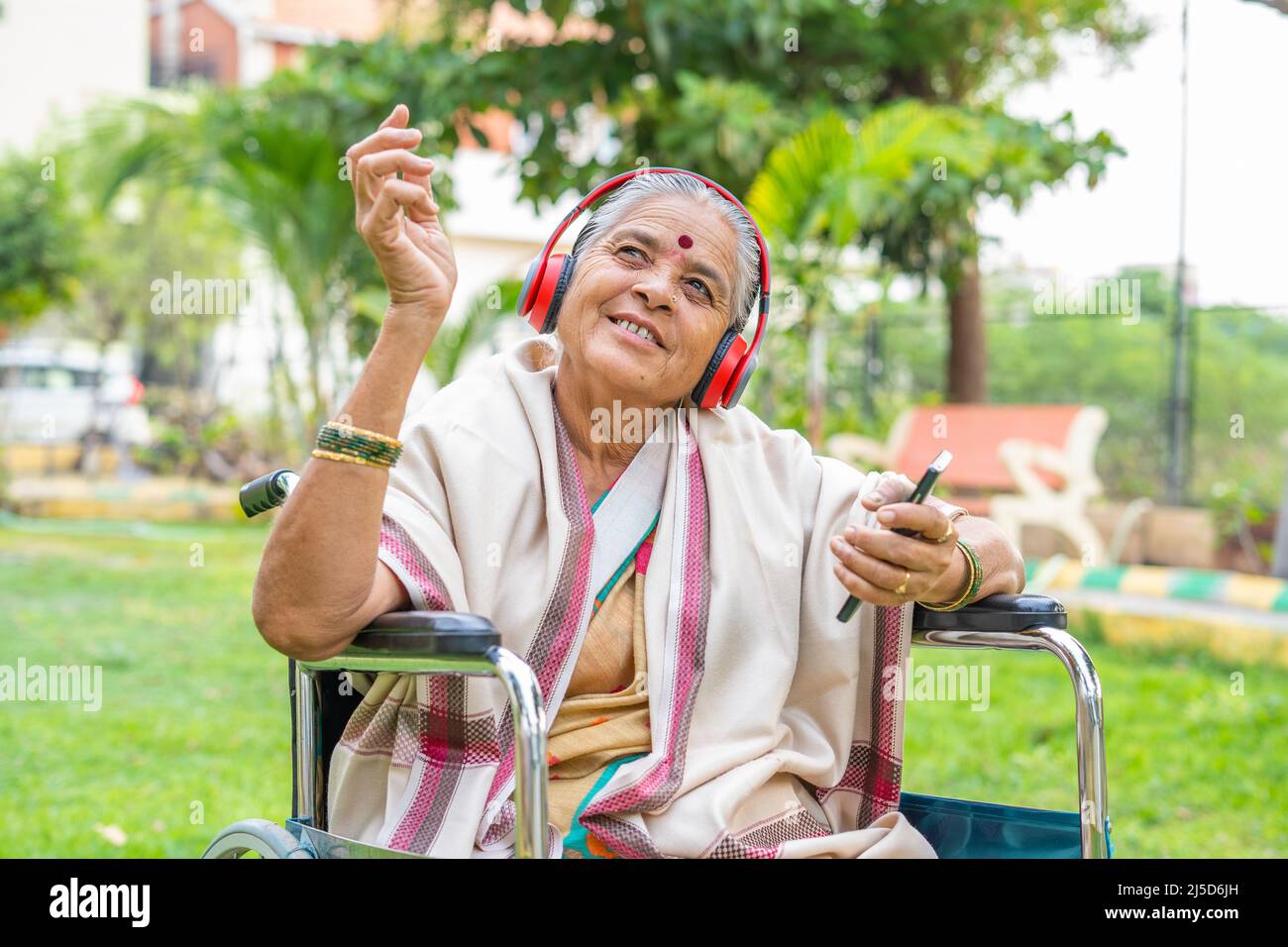 Godersi una donna anziana con disabilità ascoltare canzoni sul telefono cellulare con cuffie mentre si siede su sedia a rotelle al parco - concetto di positivo Foto Stock