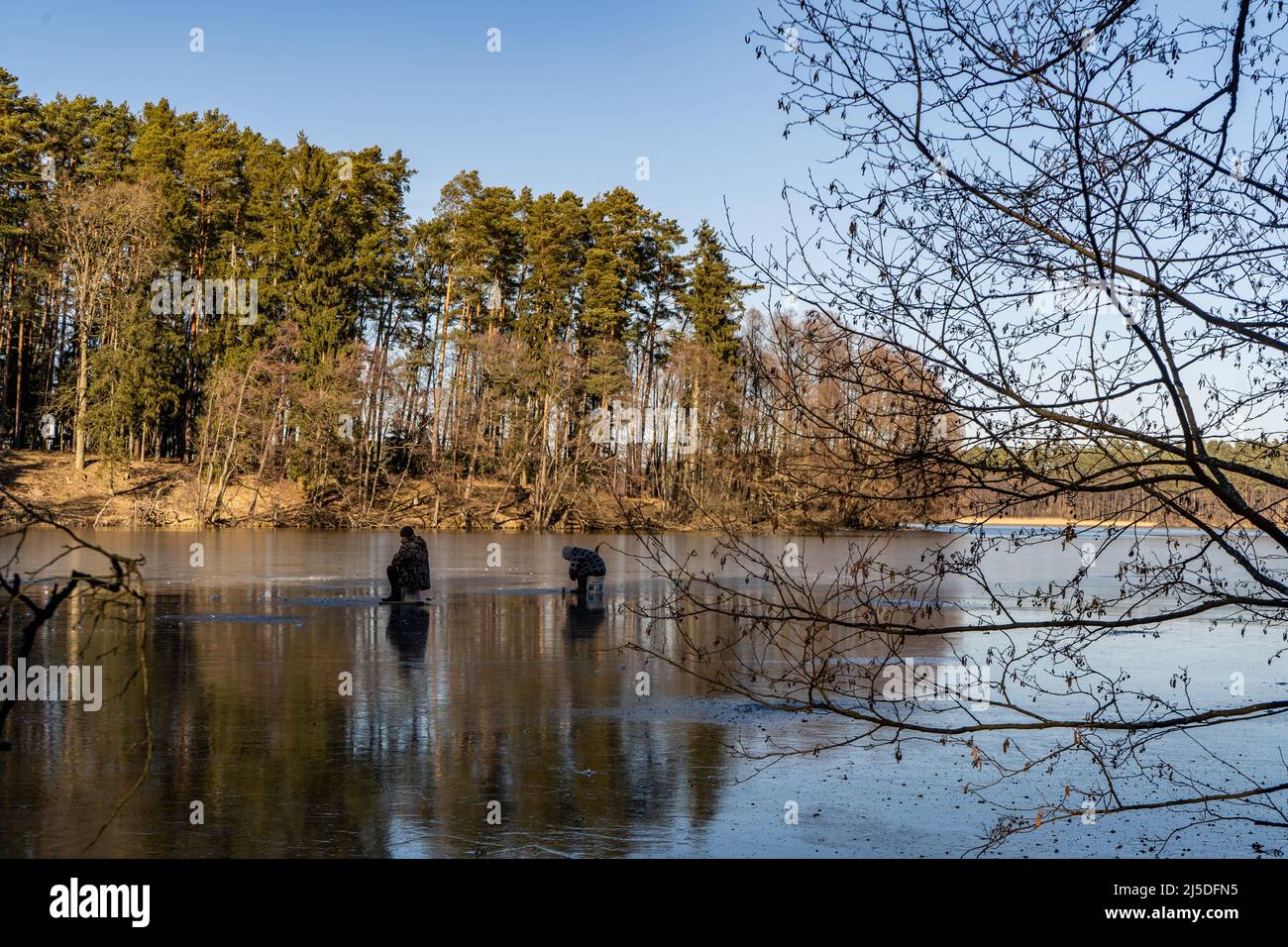 Grande angolo di cattura di due persone che si levano su un lago parzialmente ghiacciato circondato da foresta nella zona polacca vicino Drzewicz villaggio in voivodato Pomeraniano. C Foto Stock