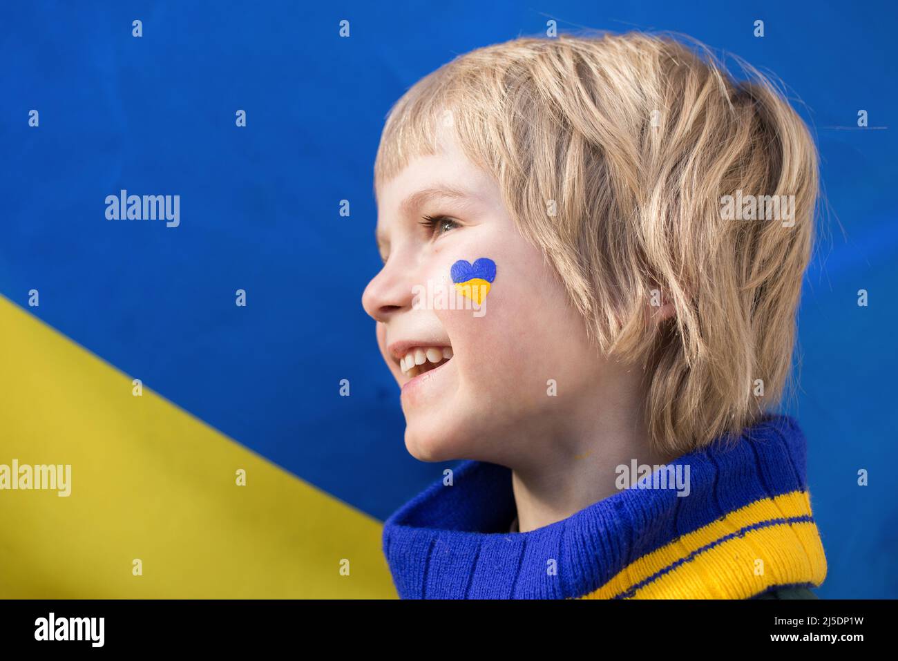 Volto di un ragazzo felice di profilo, un cuore dipinto sulla guancia con i colori giallo e blu della bandiera Ucraina. Bambini contro la guerra. Chiedere aiuto Foto Stock