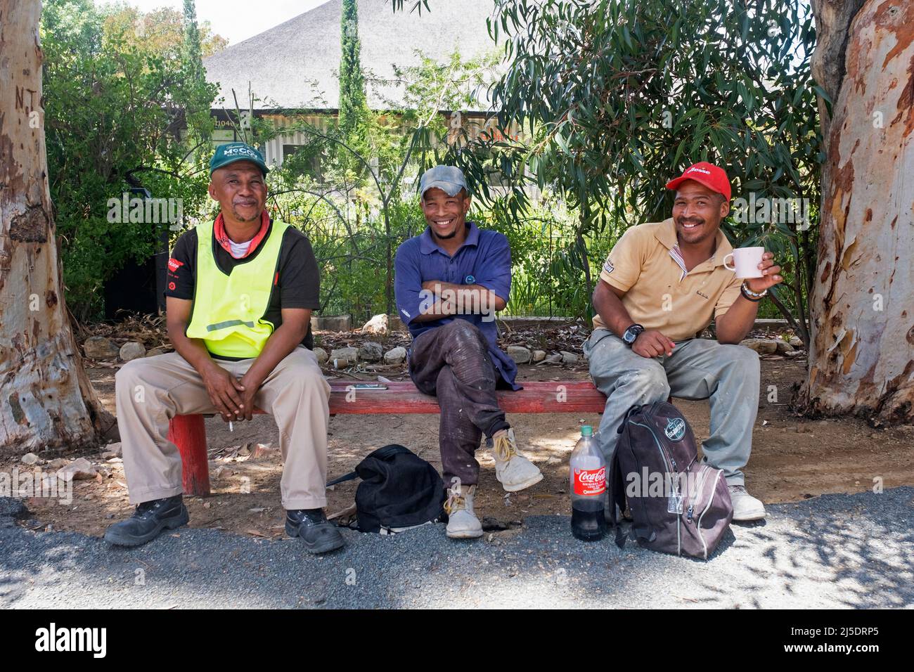 I lavoratori comunali neri felici sulla panchina ridono mentre posano durante la pausa pranzo nella città Prince Albert, Central Karoo, Western Cape, Sud Africa Foto Stock