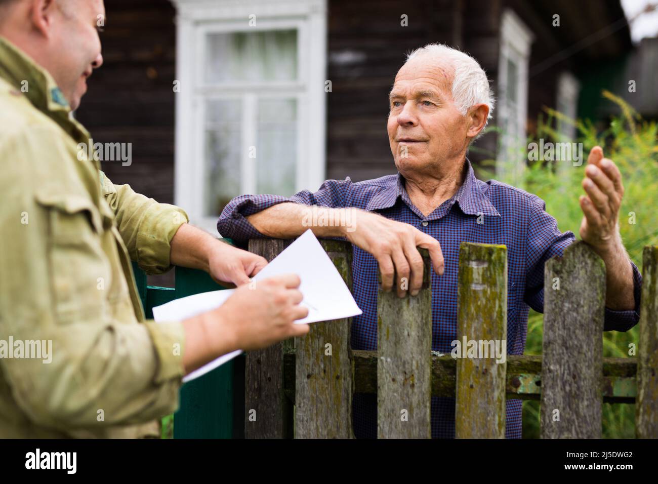 Uomo anziano che comunica con il rappresentante della compagnia di assicurazioni mentre si trova alla recinzione della sua casa di campagna Foto Stock