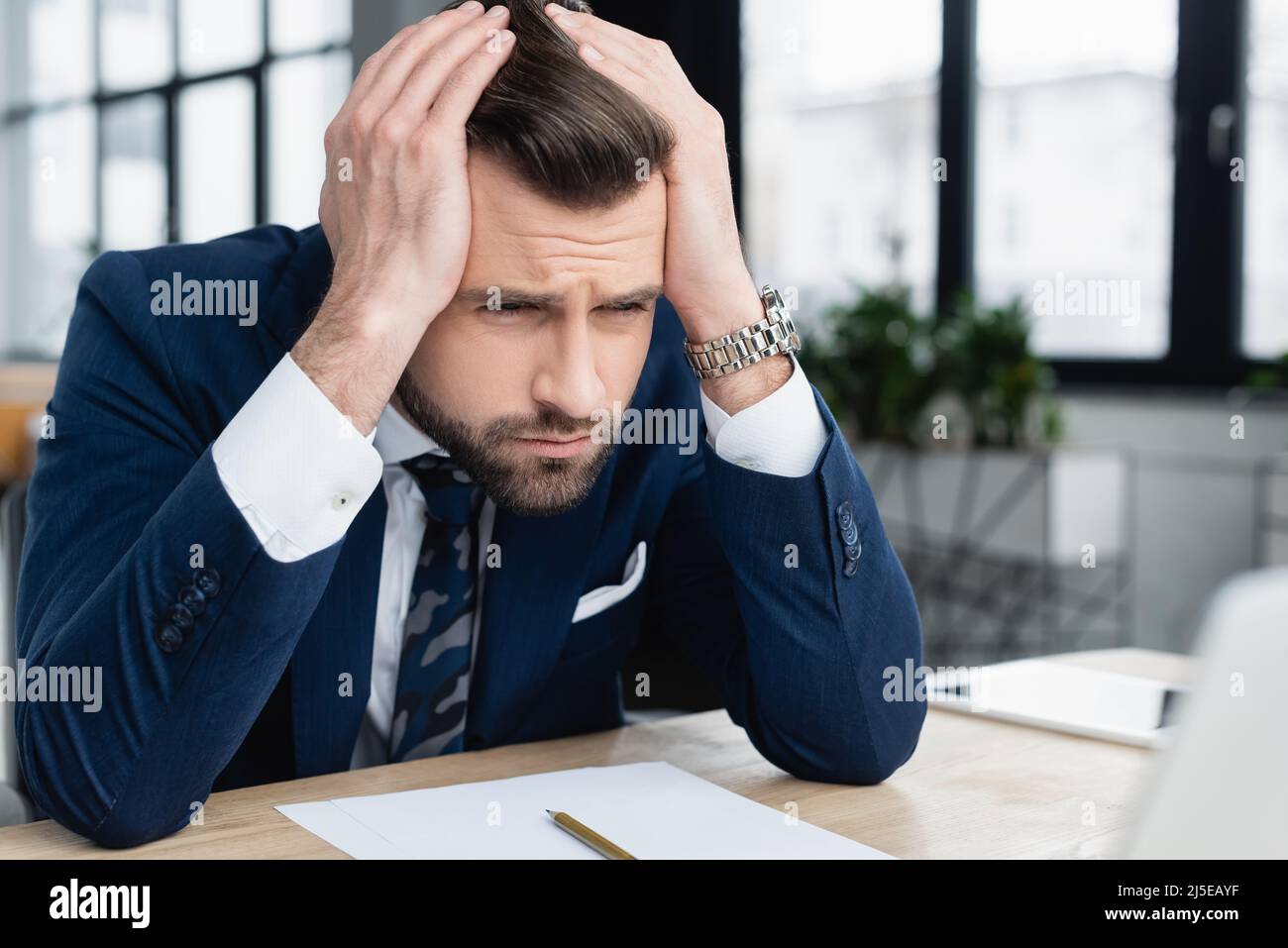 dispiacente uomo d'affari che tiene le mani sulla testa mentre si siede sul posto di lavoro Foto Stock