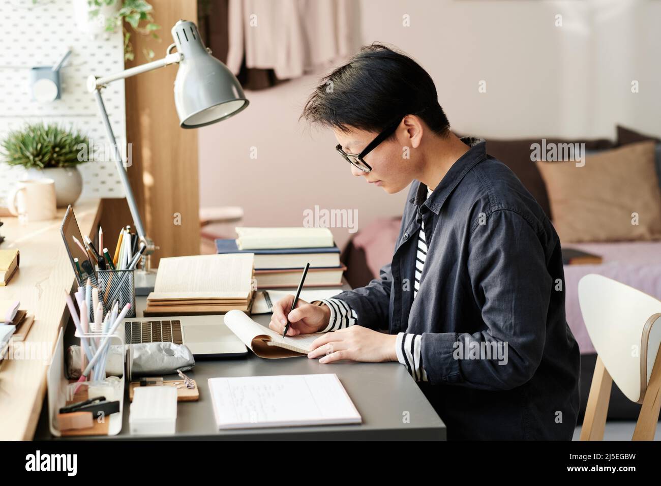 Concentrato giovane studente asiatico in occhiali seduti al tavolo in sala campus e disegno in sketchpad Foto Stock