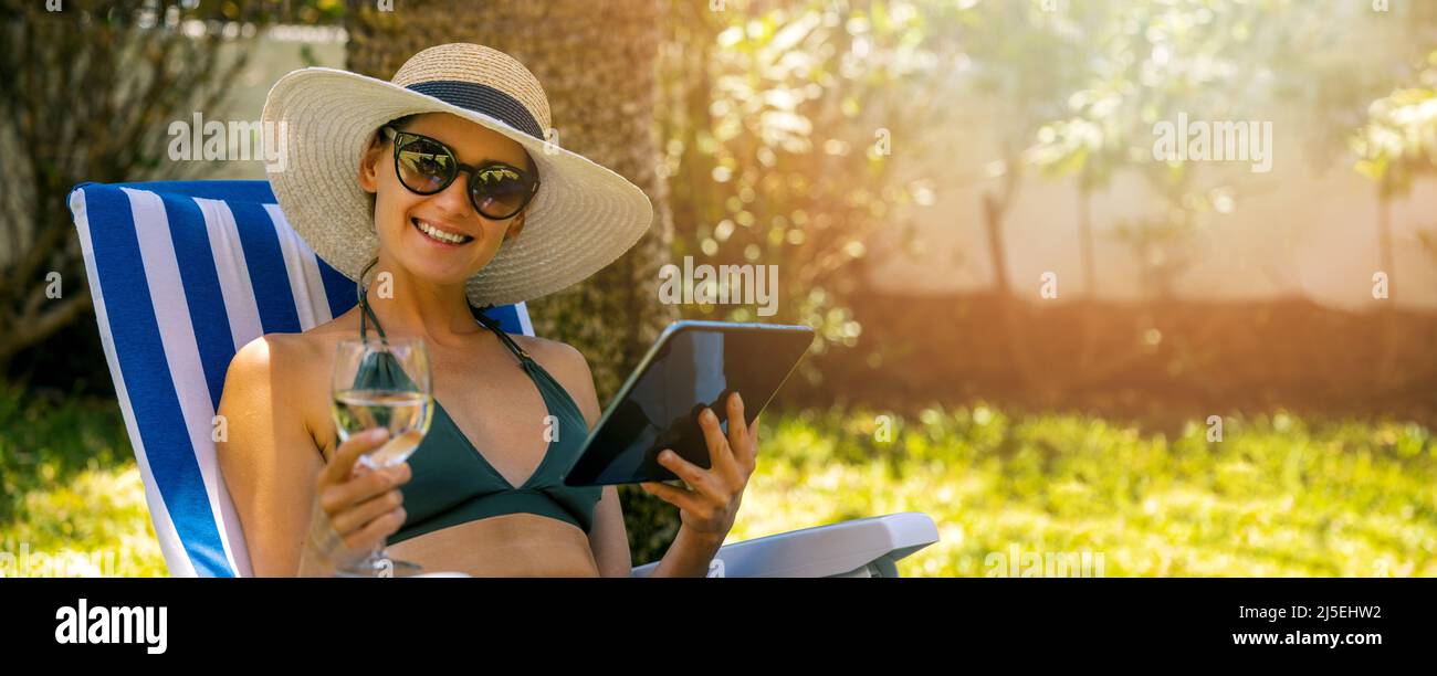 giovane donna attraente che indossa bikini rilassarsi nel lettino da sole in giardino. bere vino e utilizzare il tablet digitale. spazio copia Foto Stock