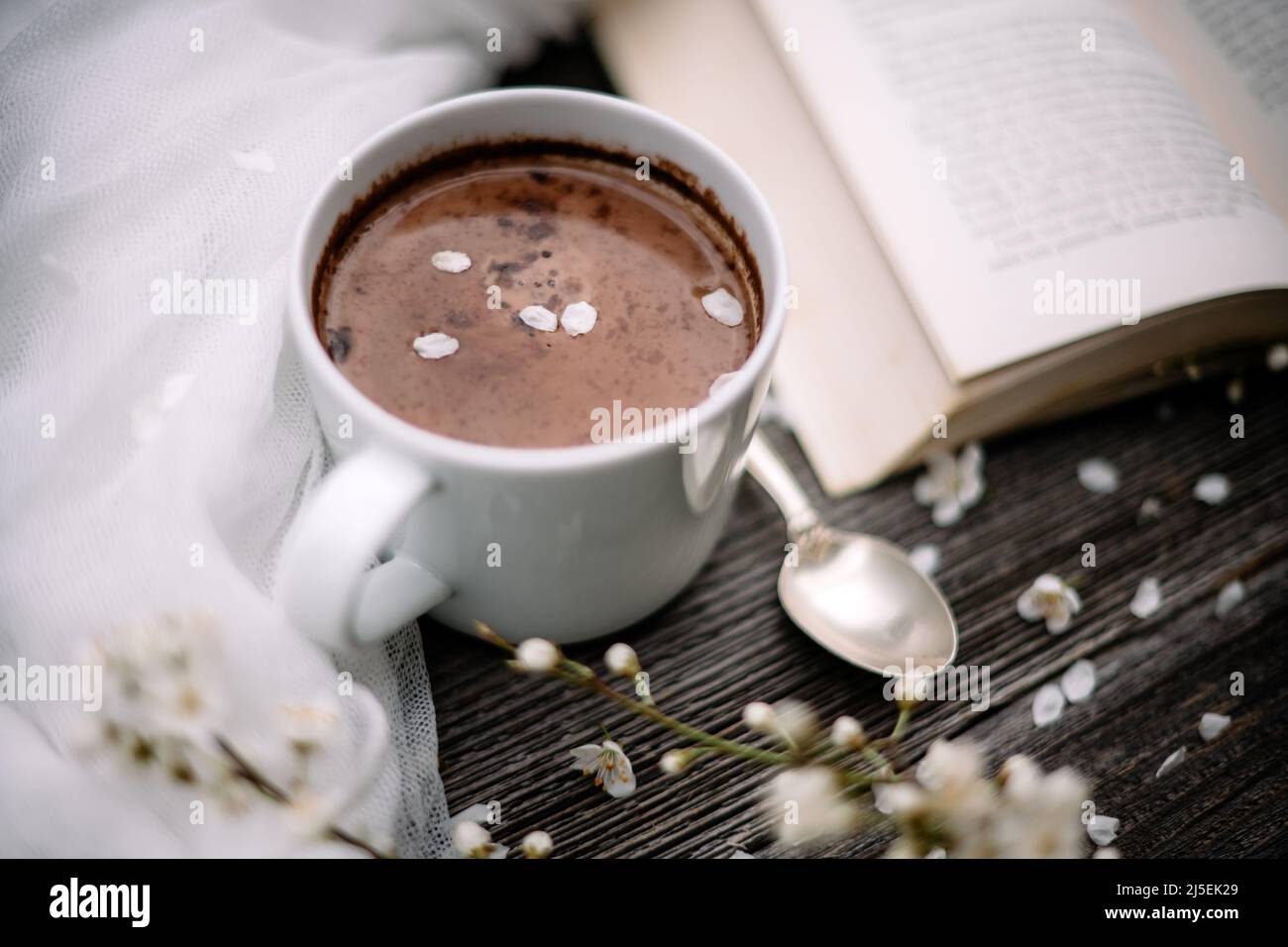 Tazza di cacao caldo al cioccolato, vecchio libro e fiori di ciliegia primaverile su sfondo di legno. Concetto di lettura Foto Stock