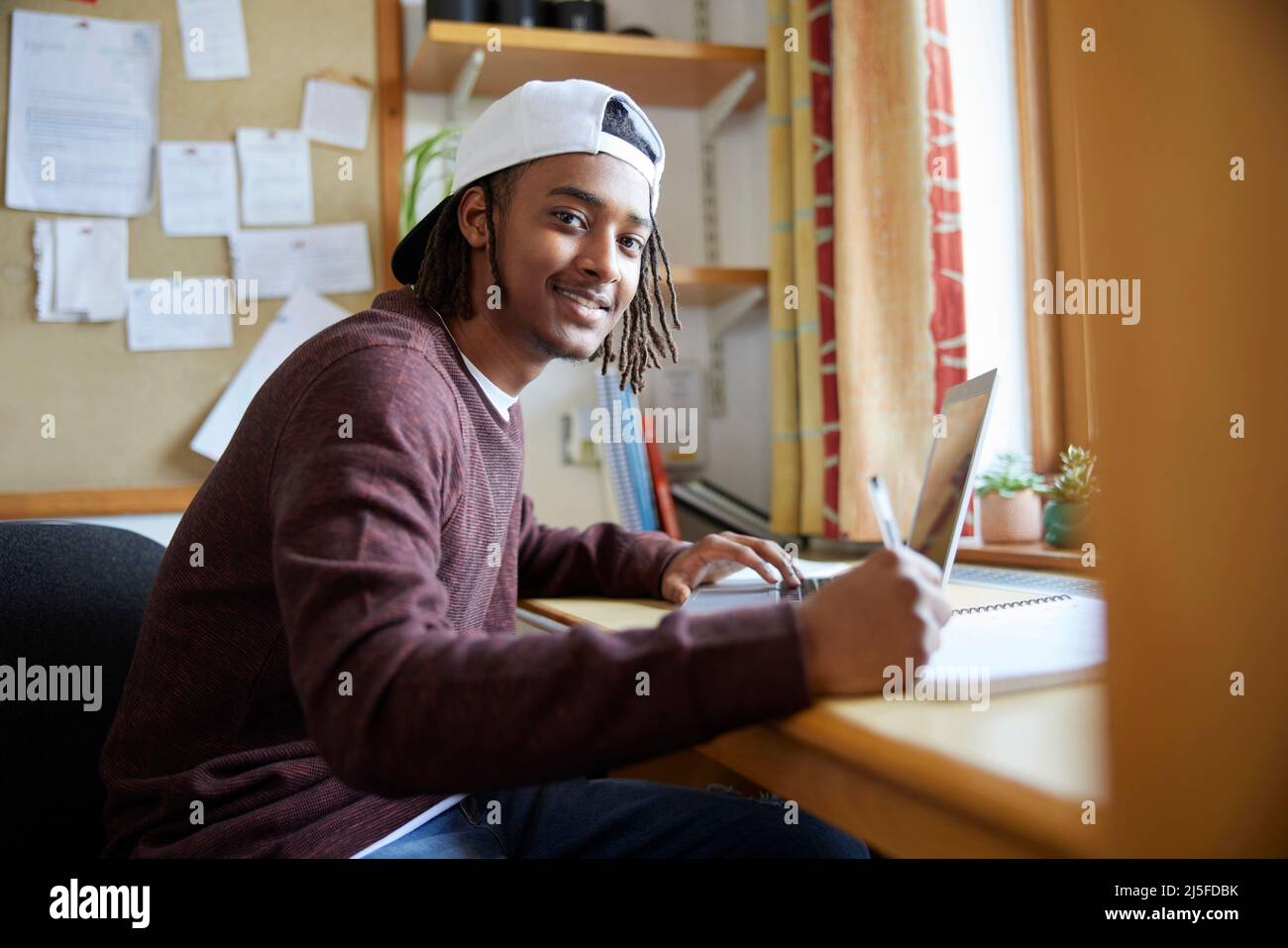 Ritratto di Male University o studente universitario indossando cappellino da baseball che studia con il laptop alla scrivania in camera Foto Stock