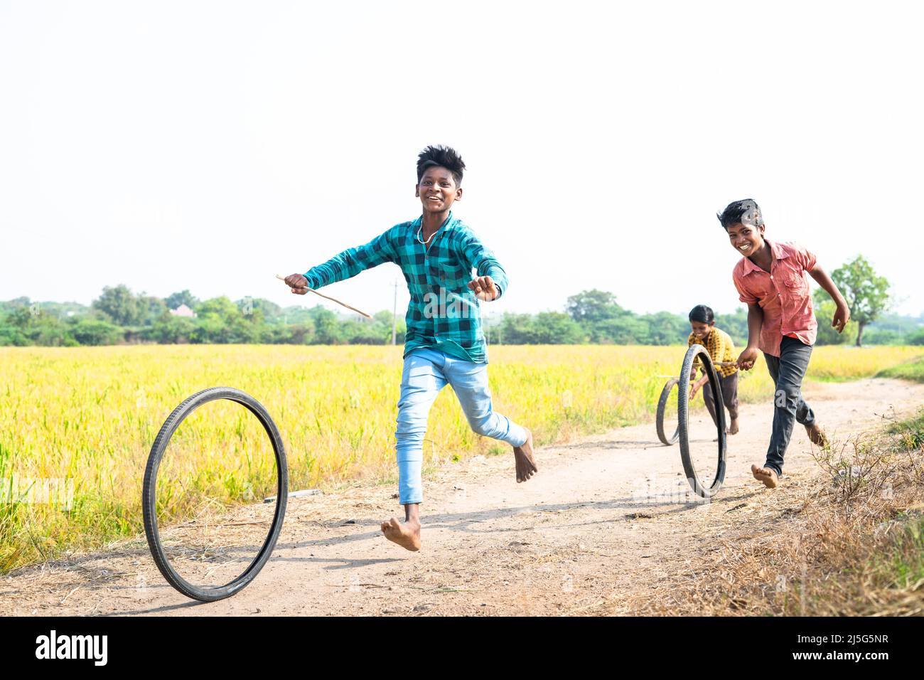 Felice allegro villaggio indiano bambini che giocano con ruota di pneumatico che rotola vicino Paddy campo rurale strada - concetto di intrattenimento, vacanze e tempo libero Foto Stock