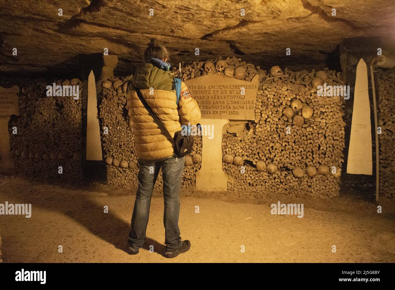 Parigi: Un visitatore di fronte a teschi, ossa e iscrizione francese con le origini dei resti nelle Catacombe di Parigi, ossario sotterraneo Foto Stock
