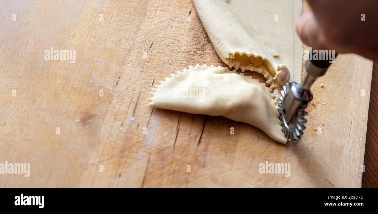 Kalitsounia preparazione tradizionale delle torte cretese. Mano femmina con una ruota di pasta frolla tagliando pasta fatta in casa farcita con formaggio di siero di latte dolce ed erbe, chiudere Foto Stock