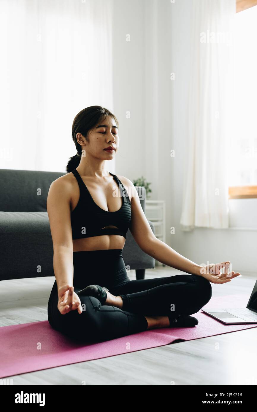 Giovane felice bella donna che pratica yoga a casa seduta in posa di loto su tappeto yoga meditando sorridente rilassato. Mentalità meditazione concetto Foto Stock