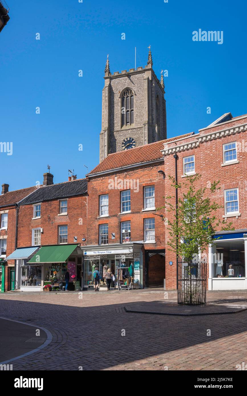 Fakenham città, vista in estate di negozi situati in Upper Market Place con la torre della chiesa parrocchiale in lontananza, Fakenham, Norfolk, Regno Unito Foto Stock