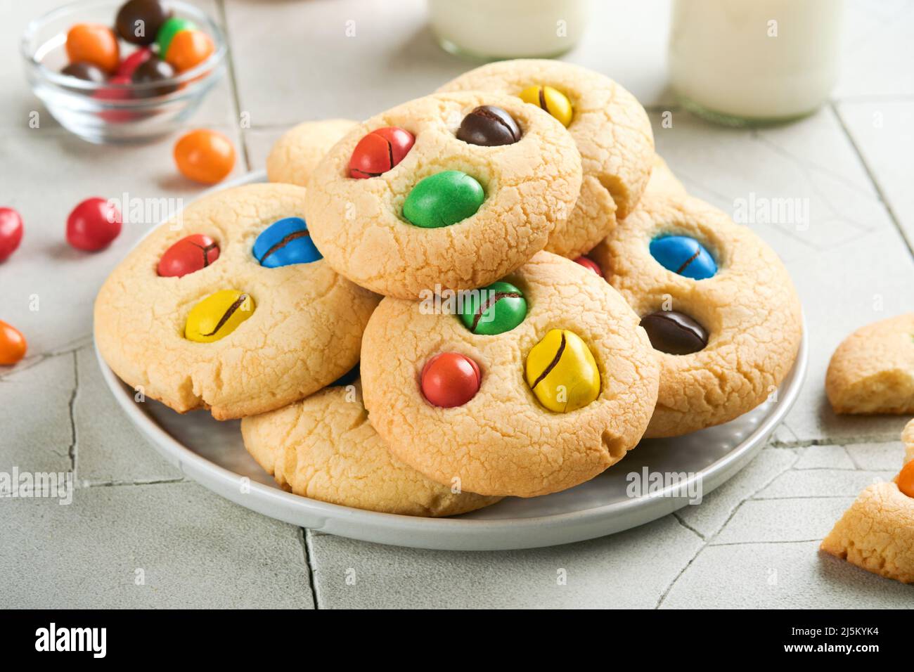 Biscotti fatti in casa con caramelle colorate al cioccolato e latte. Accatastate biscotti frollini con caramelle multicolore sul piatto con una bottiglia di latte accesa Foto Stock