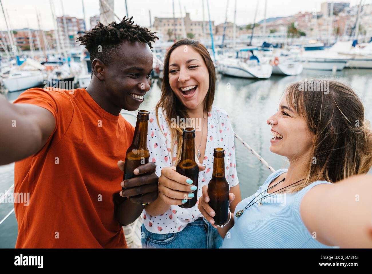 Tre amici allegri multirazziali bevono birra mentre prendono selfie in barca partito Foto Stock