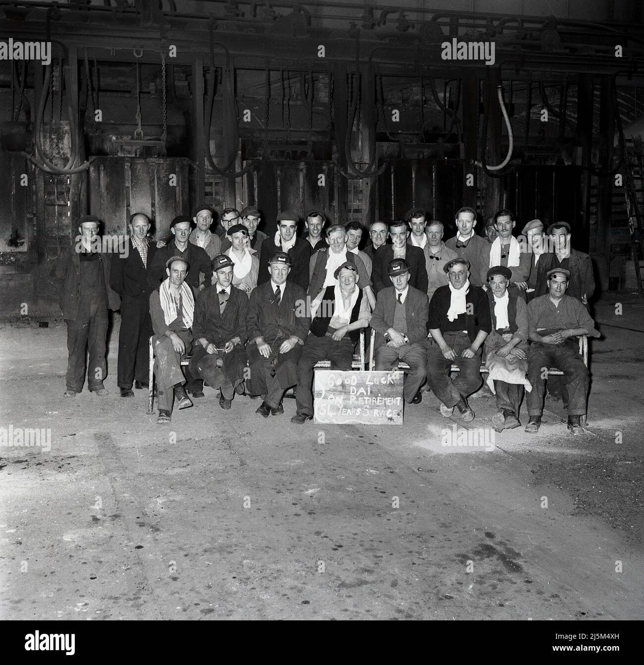 1950s, i lavoratori e i capi storici dell'acciaio si siedono insieme per una foto per comunitarizzare il ritiro di uno dei loro, dai, 34 anni un'acciaieria, Abbey Works, Margam, Port Talbot, Galles, REGNO UNITO. Segno davanti dice: 'Buona fortuna dai...su pensionamento....54 anni di servizio. Foto Stock