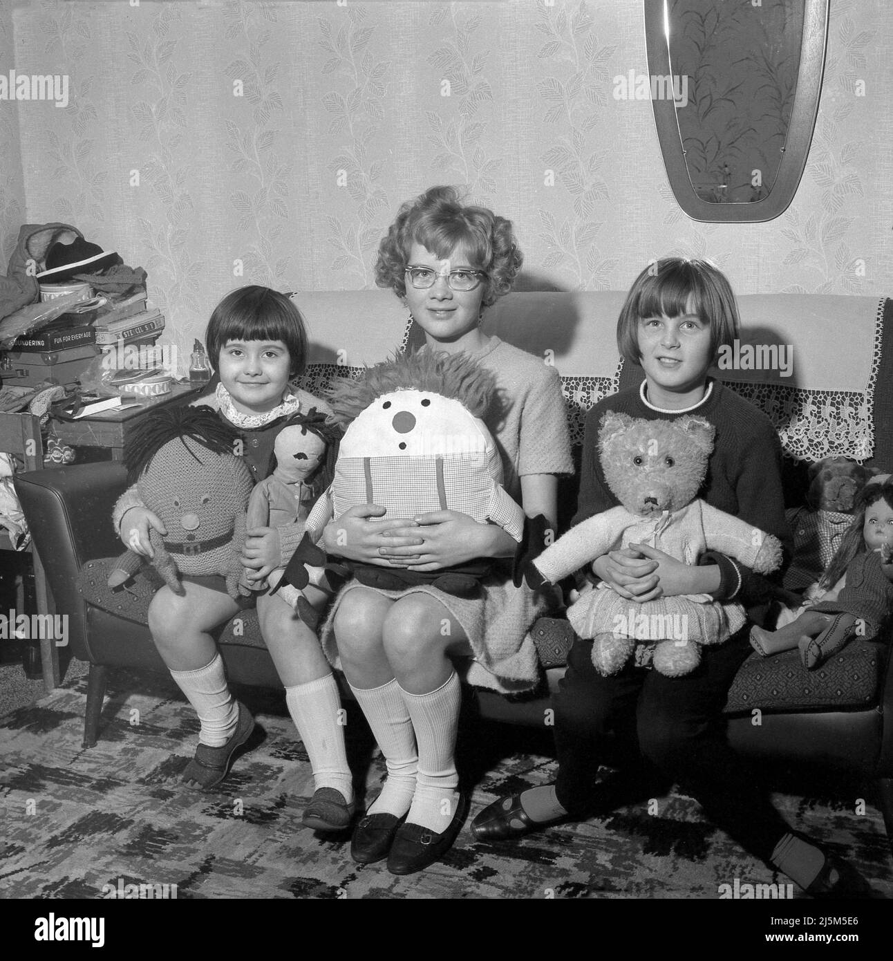 1966, storica, tre giovani ragazze che si siedono insieme su un divano in una stanza anteriore che tiene i loro giocattoli molli, ragdoll e orsacchiotti. Foto Stock