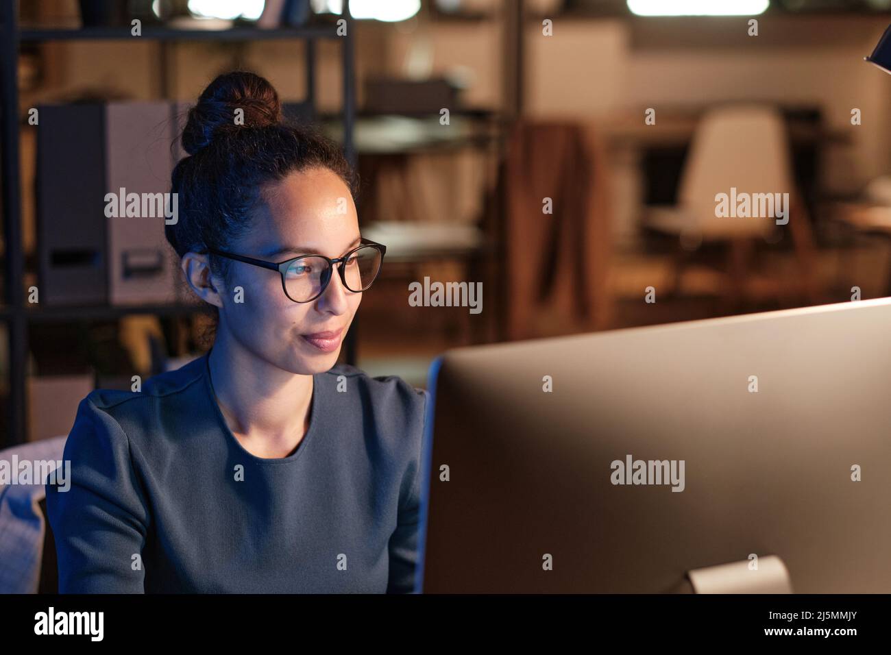 Soddisfare giovane donna biraciale impiegato in ufficio in occhiali utilizzando il computer durante il lavoro di notte Foto Stock